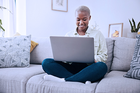 Smiling black woman on sofa with laptop on crossed legs